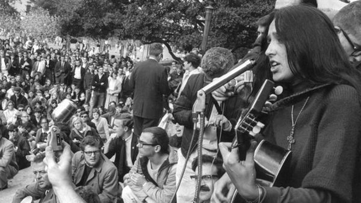 Rechts im Vordergrund steht eine Sängerin mit Gitarre. Im Hintergrund sind viele sitzende und stehende Menschen zu sehen. | Bildquelle: dpa Rechts im Vordergrund steht eine Sängerin mit Gitarre. Im Hintergrund sind viele sitzende und stehende Menschen zu sehen.