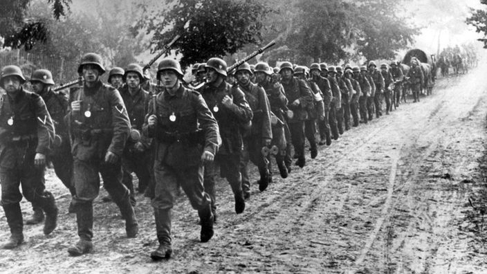 In einer langen Reihe marschieren deutsche Soldaten entlang eines Bachlaufes. Sie sind bewaffnet und tragen Rucksäcke.