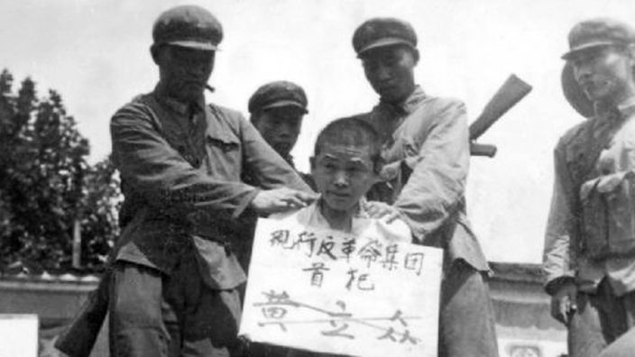 Schwarzweiß-Foto: Armeemitglieder mit einem Gefangenen, der vor ihnen kniet und ein Schild um den Hals hat