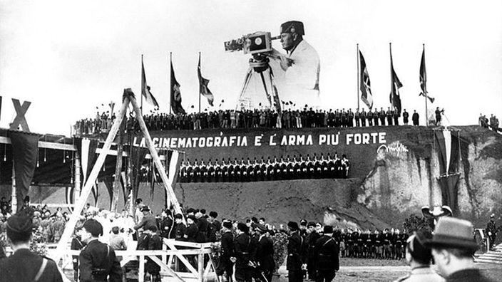 Mussolini-Konterfei mit Filmkamera über einer Menschenmenge