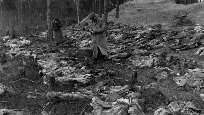 Schwarzweiß-Foto: Identifizierung der Toten, nach der Entdeckung der Massengräber von Katyn 1943. Zwei Männer stehen mit Schreibblöcken in einem Meer exhumierter Leichen.