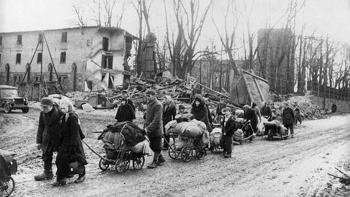 Eine Gruppe von Flüchtlingen zieht 1944 ihre Habseligkeiten auf Leiterwagen durch eine zerstörte Ortschaft in Ostpreußen.