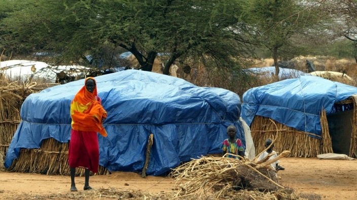 Eine Frau steht in einem sudanesischen Flüchtlingslager vor Strohhütten, die mit Plastikplanen abgedeckt sind.