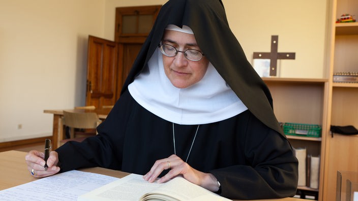 Eine Nonne sitzt an einem Schreibtisch, liest aus einem Buch und hält einen Stift in der rechten Hand.