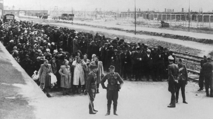 Neu angekommene Häftlinge haben auf der Todesrampe im KZ Auschwitz Aufstellung genommen. Links Frauen und Kinder, rechts die Männer. Im Hintergrund sind Gleisanlagen und Gebäude hinter Stacheldraht zu sehen.