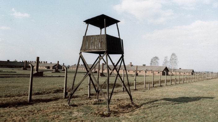 Ein hölzerner Wachturm vor einem Stacheldrahtzaun. Im Hintergrund stehen mehrere flache lange Ziegelbauten. | Bildquelle: akg Ein hölzerner Wachturm vor einem Stacheldrahtzaun. Im Hintergrund stehen mehrere flache lange Ziegelbauten.