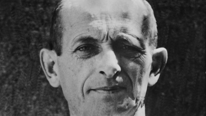 Die Schwarz-Weiß-Fotografie zeigt Adolf Eichmann im weißen Hemd. Er guckt grimmig in die Kamera. Die Aufnahme stammt aus dem Mai 1960.