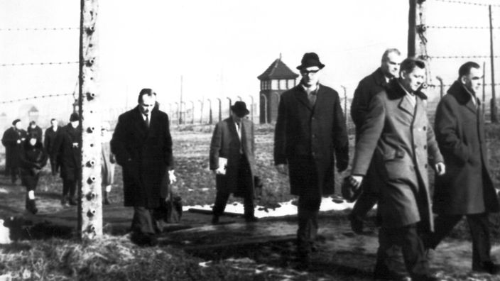 Mehrere Männer in langen Mänteln gehen auf diesem Schwarz-Weiß-Foto durch einen geöffneten Stacheldrahtzaun im Lager Auschwitz.