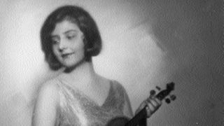 Schwarzweiß-Foto einer jungen Frau. Sie trägt ein langes Kleid, hält in der linken Hand eine Geige, in der rechten einen Geigenbogen und blickt lächelnd nach unten.