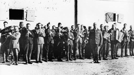 Schwarzweiß-Foto einer Gruppe Männer, die vor einer Gebäudewand stehen. Einige spielen Blas-, andere Streichinstrumente, zwei Männer spielen Akkordeon.