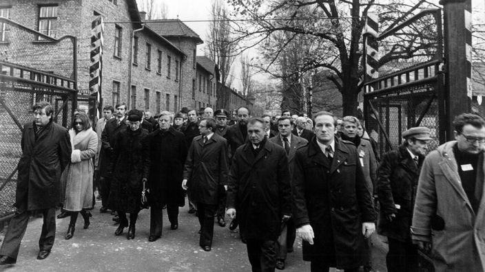 Der ehemalige Bundeskanzler Helmut Schmidt in einem Tross von Menschen am Eingang des Konzentrationslager Auschwitz. Über dem Eingangstor stehen die berühmten Worte 'Arbeit macht frei'.