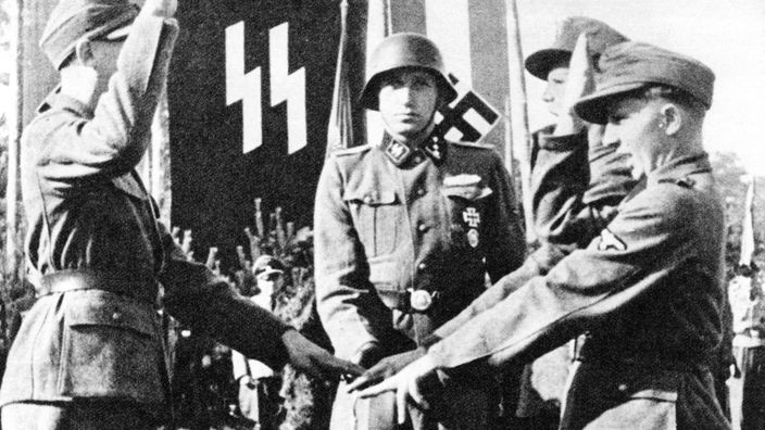Ein junger Mann in Uniform steht vor einer SS-Fahne und wird vereidigt.
