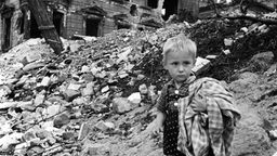 Ein kleiner Junge mit einer Decke im Arm auf einem Trümmerberg vor einem zerbombten Haus