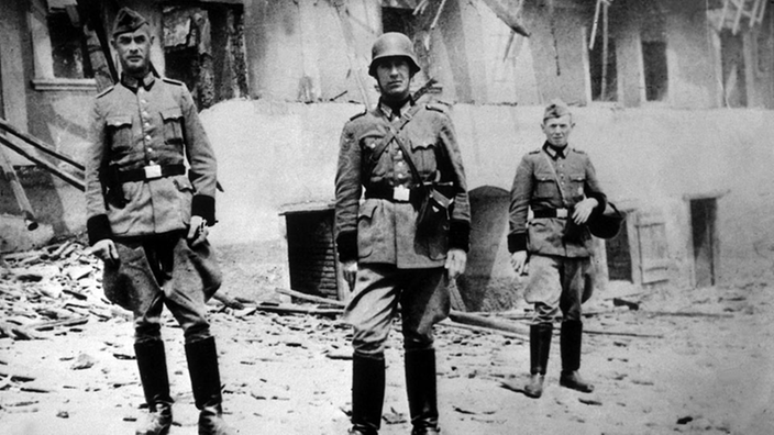 Schwarzweiß-Aufnahme: Drei Soldaten der Wehrmacht stehen vor Häuserruinen im Dorf Lidice.