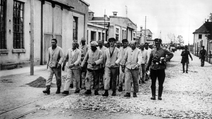 Schwarzweiß-Bild: Häftlinge marschieren in Fünfer-Reihen, daneben ein Aufseher.