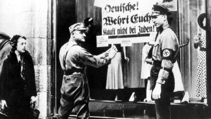 Schwarzweiß-Bild: SA-Männer kleben ein Plakat, auf dem steht 'Deutsche! Wehrt Euch! Kauft nicht bei Juden!' an das Schaufenster eines jüdischen Geschäftes.