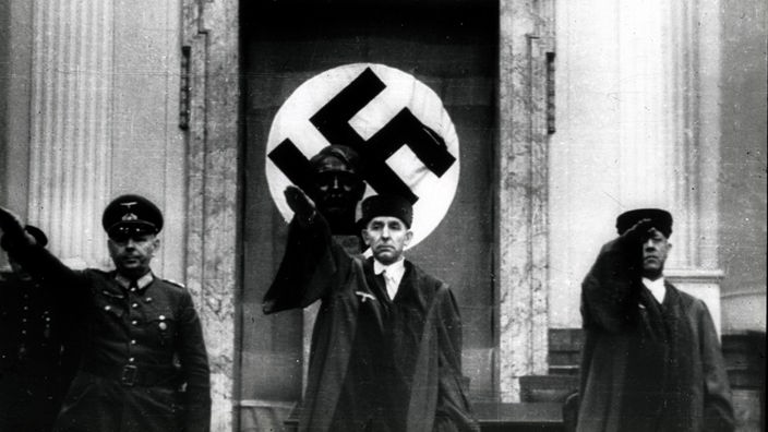 Roland Freisler eröffnet zwischen zwei anderen Männern stehend ein Verfahren des Volksgerichtshofes. Alle drei haben die Hand zum Hitlergruß erhoben, hinter ihnen hängt Flagge mit dem Hakenkreuz und steht eine Hitler-Büste.