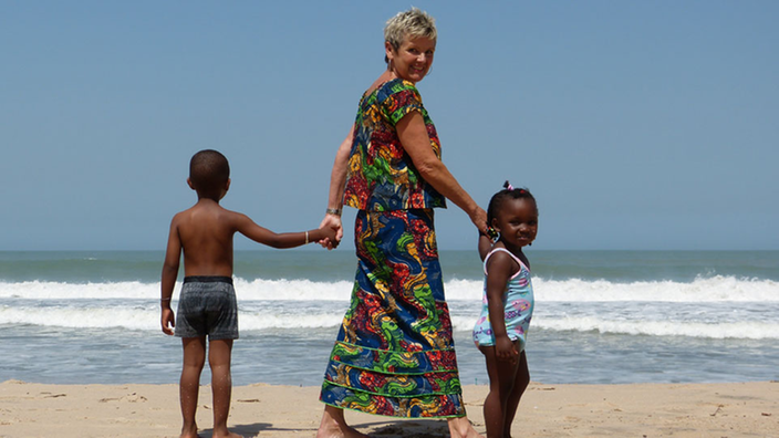 Eine ältere Frau steht am Strand und hält zwei kleine Kinder an den Händen.