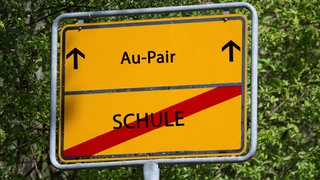 Auf einem Schild steht das Wort "Schule" durchgestrichen, darüber das Wort "Au-Pair"