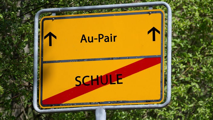 Auf einem Schild steht das Wort "Schule" durchgestrichen, darüber das Wort "Au-Pair"
