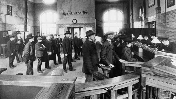 Menschen stehen in Schlangen an Ticketschaltern im Hamburger Hafen, alte Schwarzweiß-Aufnahme. | Bildquelle: Picture Alliance Menschen stehen in Schlangen an Ticketschaltern im Hamburger Hafen, alte Schwarzweiß-Aufnahme.