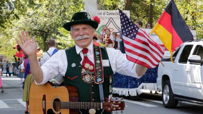 Ein Mann in Lederhose und Trachtenhut hält bei der Steubenparade in New York eine deutsche und eine US-Flagge