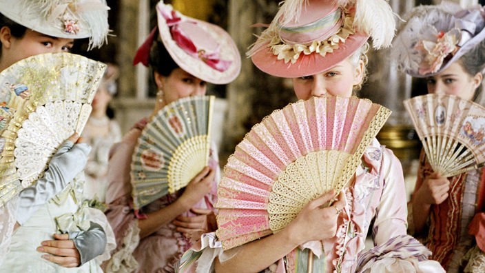 Schauspielerin Kirsten Dunst und weitere halten sich in prunkvollen Kostümen bunte Fächer vors Gesicht.