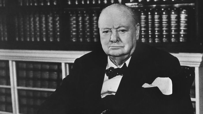 Winston Churchill mit braunem Wintermantel, schwarzem Hut und einer Zigarre im Mund.
