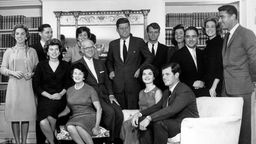 Der Kennedy-Clan mit Schwiegersöhnen und -töchtern auf einem Gruppenbild im Haus der Familie. Einige Personen sitzen, andere stehen vor einer Bücherwand