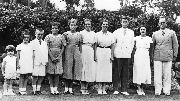 Schwarzweiß-Gruppenbild der Kennedys. Die acht Kinder sowie ihre Eltern stehen in einem Garten und lächeln in die Kamera.