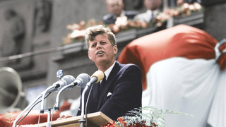 'JFK' steht vor drei Mikrofonen. Im Hindergrund weht eine Fahne und drei Männer stehen auf einem Balkon.