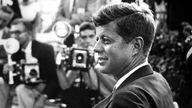 John F. Kennedy sitzt im Vordergrund und wird von mehreren Fotografen aufgenommen.
