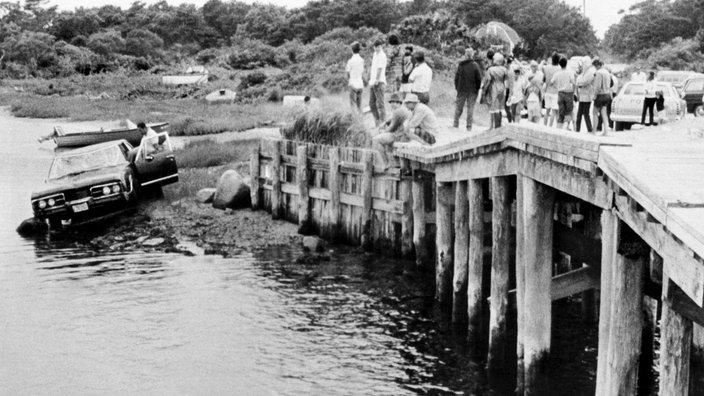 Eine Gruppe Menschen schaut von einer Brücke auf ein Auto hinunter, das im Wasser liegt.