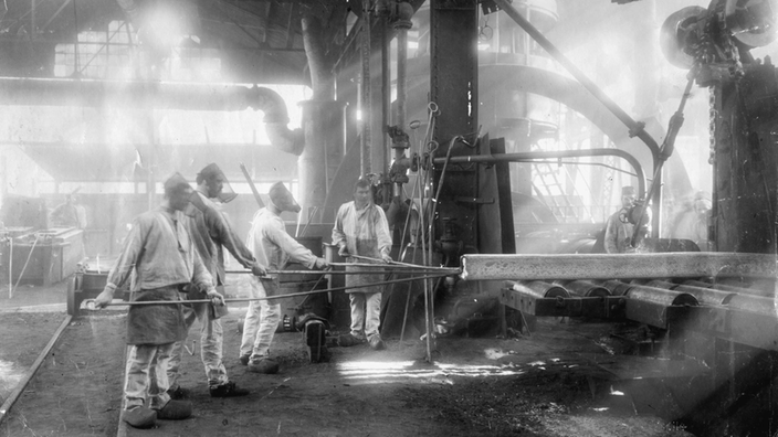 Innenaufnahme einer Werkshalle bei Krupp, in der Arbeiter eine Schiene bearbeiten.