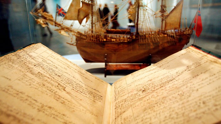 Das Logbuch von James Cook liegt in einer Glasvitrine. Dahinter erkennt man ein Schiffsmodell. 