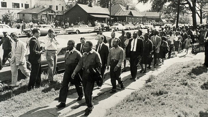 Schwarzweiß-Aufnahme: Martin Luther King marschiert zusammen mit anderen Schwarzen durch einen amerikanischen Vorort