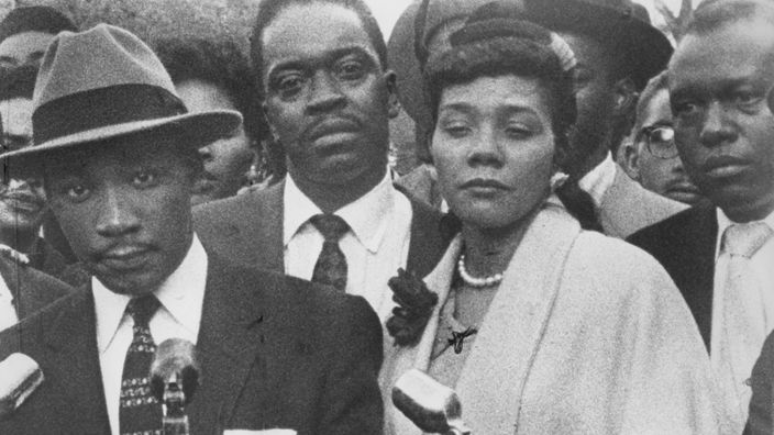 Schwarzweiß-Foto: Martin Luther King vor Mikrofonen, daneben seine Ehefrau und einige Vertraute