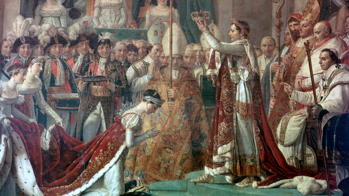 Joséphine kniet bei der Krönungszeremonie vor Napoleon, der die Krone in seinen Händen hält.