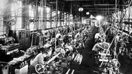 Das Schwarzweiß-Foto einer großen Fabrikhalle im 19. Jahrhundert. Dicht gedrängt steht Arbeiter an Arbeiter.