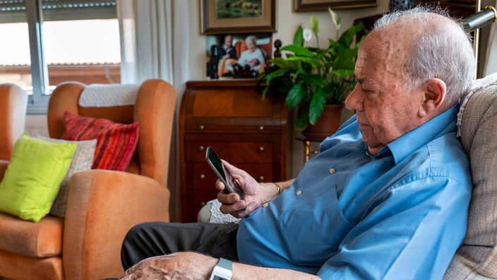 Ein alter Mann sitzt Zuhause im Sessel mit einem Smartphone in der Hand.