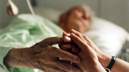 Ein alter Mensch in einem Klinikbett; seine Hände werden von zwei Händen eines jungen Menschen gehalten.