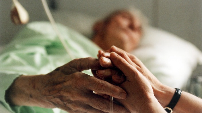 Ein alter Mensch in einem Klinikbett; seine Hände werden von zwei Händen eines jungen Menschen gehalten. | Bildquelle: SWR/Imago Ein alter Mensch in einem Klinikbett; seine Hände werden von zwei Händen eines jungen Menschen gehalten.