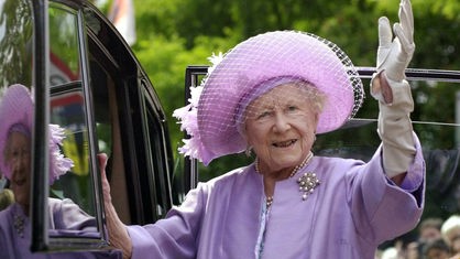 Das Foto zeigt eine Großaufnahme der lächelnden Königinmutter Elizabeth, die aus einem Auto heraus winkt. Sie hat einen lilafarbenen Hut mit Schleier auf und trägt ein gleichfarbiges Kostüm.