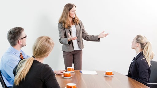 Eine Chefin steht gestikulierend am Konferenztisch, um sie herum sitzen zwei Frauen und ein Mann