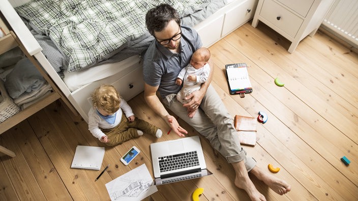 Ein Mann sitzt auf dem Boden im Kinderzimmer, mit Laptop auf dem Schoß und Baby im Arm