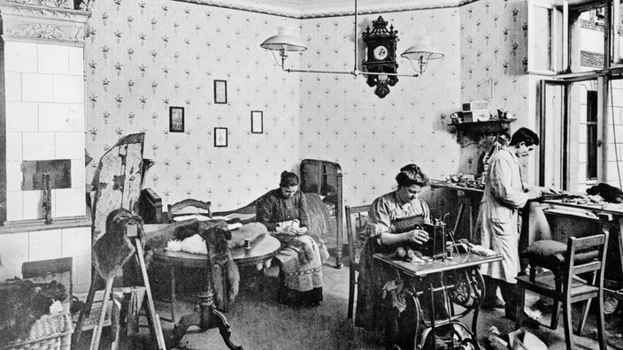 Schwarz-Weiß-Fotografie eines Wohnraums, in dem zwei Frauen und ein Mann Näharbeiten ausführen.