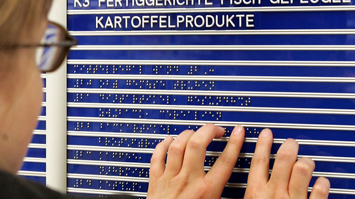 Eine Frau ertastet die Braille-Schrift auf einer blauen Tafel. Auf der Tafel steht, wo sich die verschiedenen Waren in einem Supermarkt befinden.