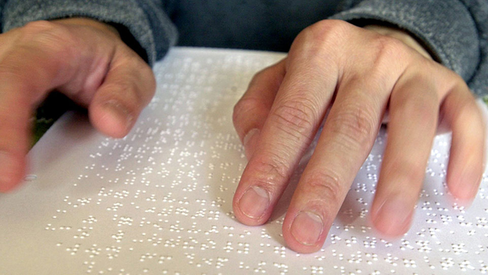 Zwei Hände ertasten die erhabenen Punkte auf einem Dokument, das in Braille-Schrift verfasst ist.