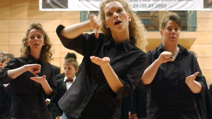 Das Foto zeigt drei schwarz bekleidete junge Mädchen, die Gebärden mit den Händen machen. Im Vordergrund ist ein blondes Mädchen, das den Mund geöffnet hat, als ob es singen würde. Über ihnen hängt ein Plakat, auf dem steht &#8218;Tage der Laienmusik&#8217;.