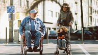Ein älterer Mann im Rollstuhl mit einem lächelnd schauendem Kind im Kinderwagen neben sich